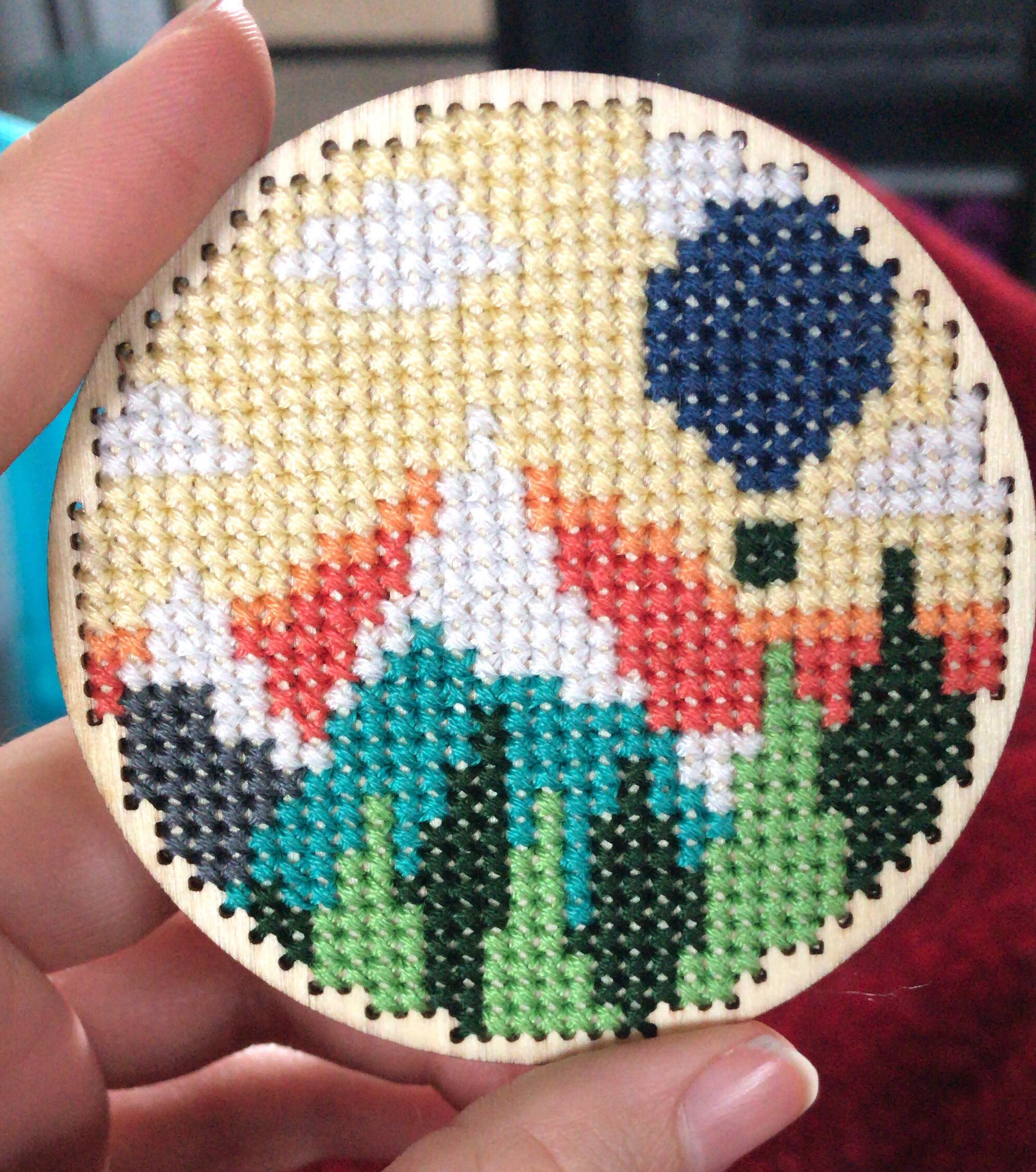 A cross stitch on a circular piece of wood of a mountain landscape with a sun